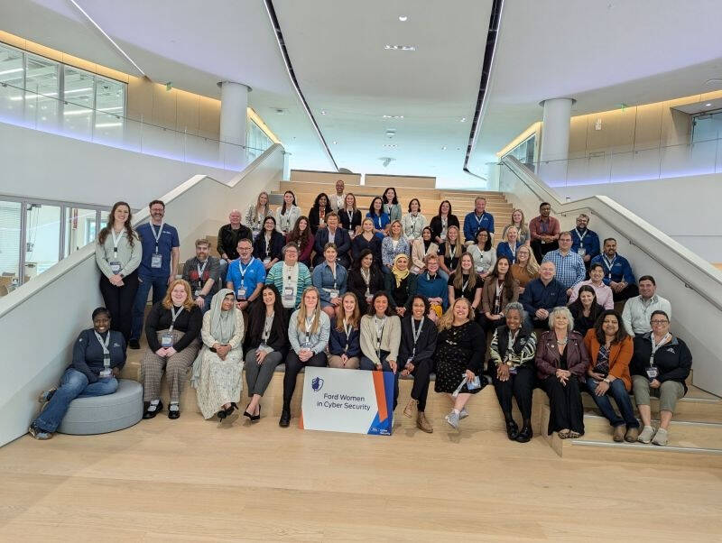 Alt text: A large group of diverse individuals posing on a wide indoor staircase in a modern building. The group includes mostly women, with a few men, all wearing name badges. A sign in front reads "Ford Women in Cyber Security."